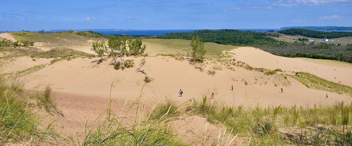 Sand dunes, grassy areas, and a distant view of the ocean under a blue sky.