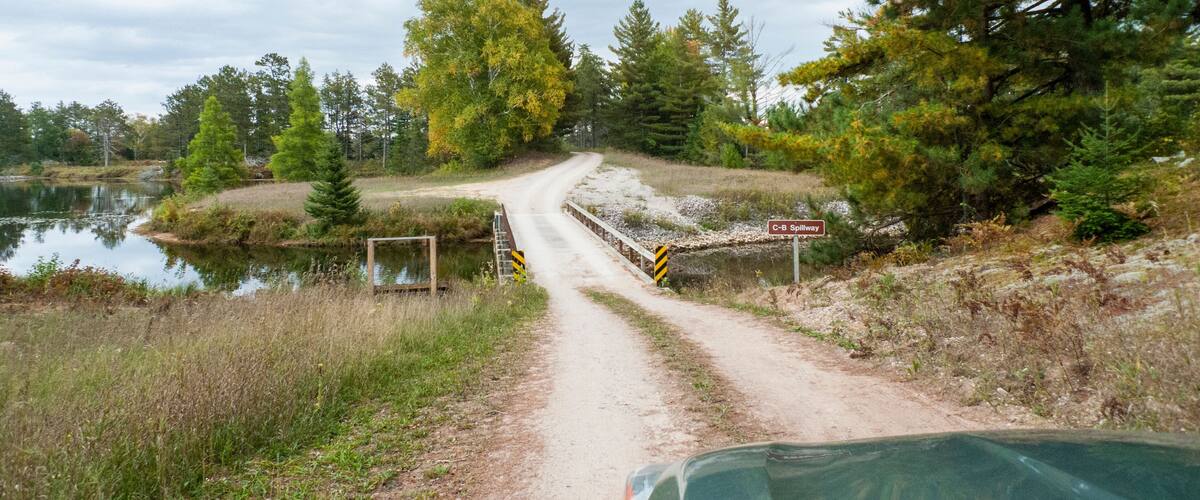 Een zandweg die leidt naar een overstortbaksysteem aan een meer, met bomen en een bewolkte lucht.
