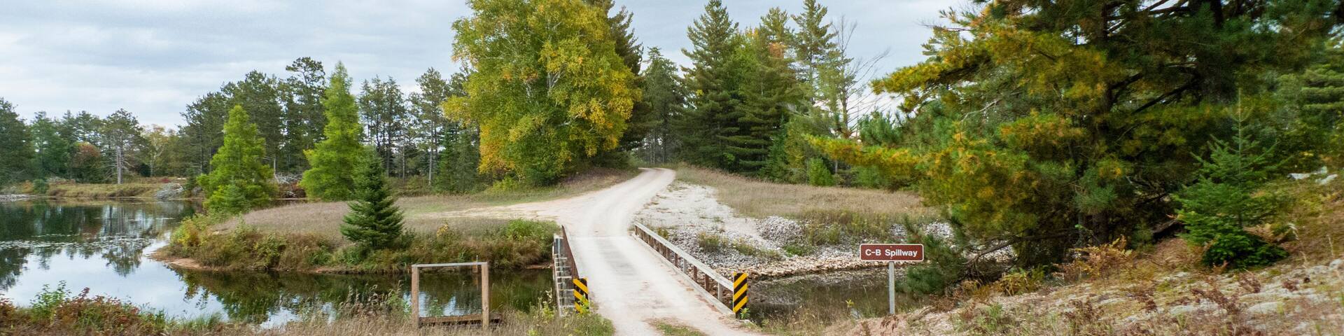 A dirt road leading to a spillway by a lake with trees and a cloudy sky.
