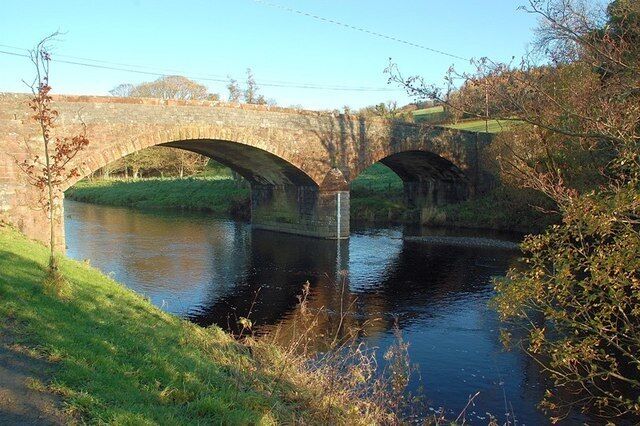 A stone bridge over a calm river with grassy banks and trees.