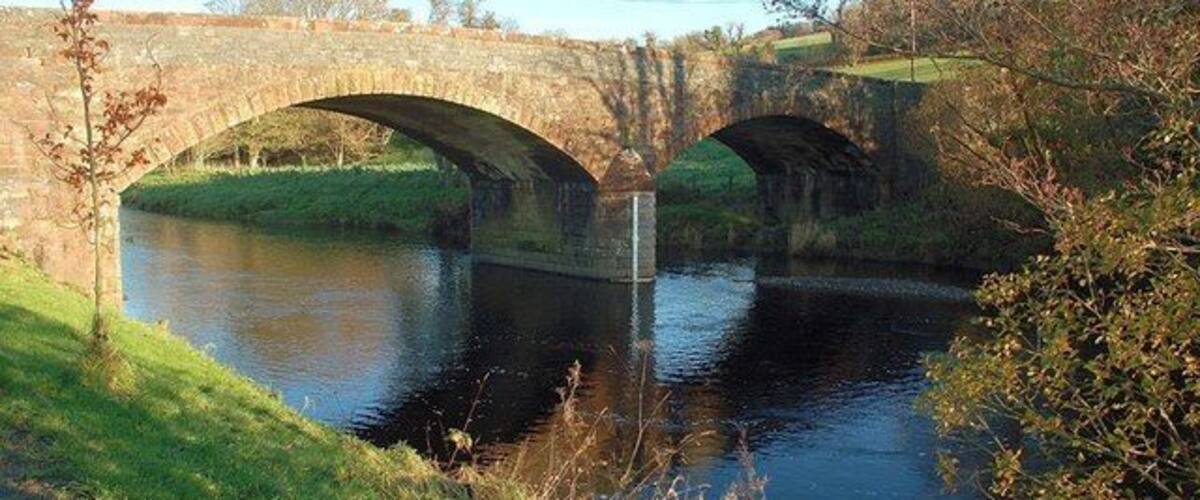 A stone bridge over a calm river with grassy banks and trees.