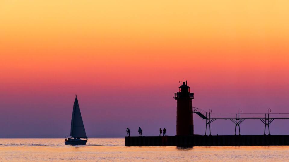 A sailboat on the water, a lighthouse, and a pier with people standing on it.