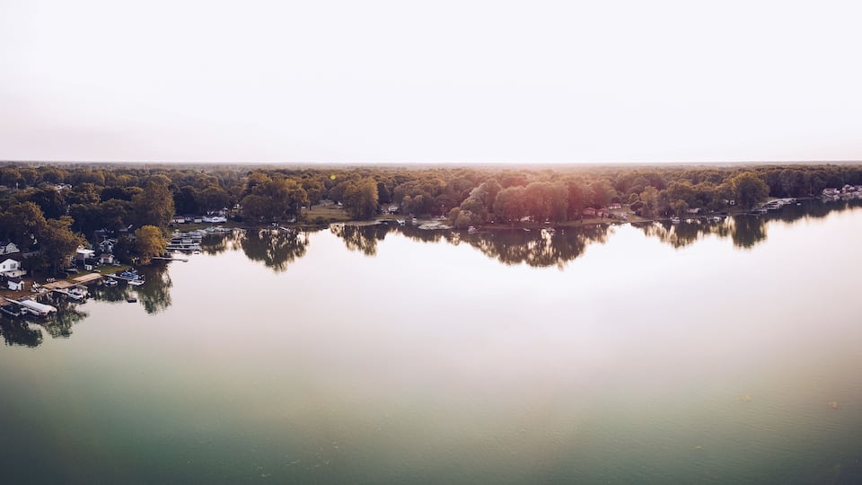 A calm lake with reflections of trees and houses, surrounded by a forested area.