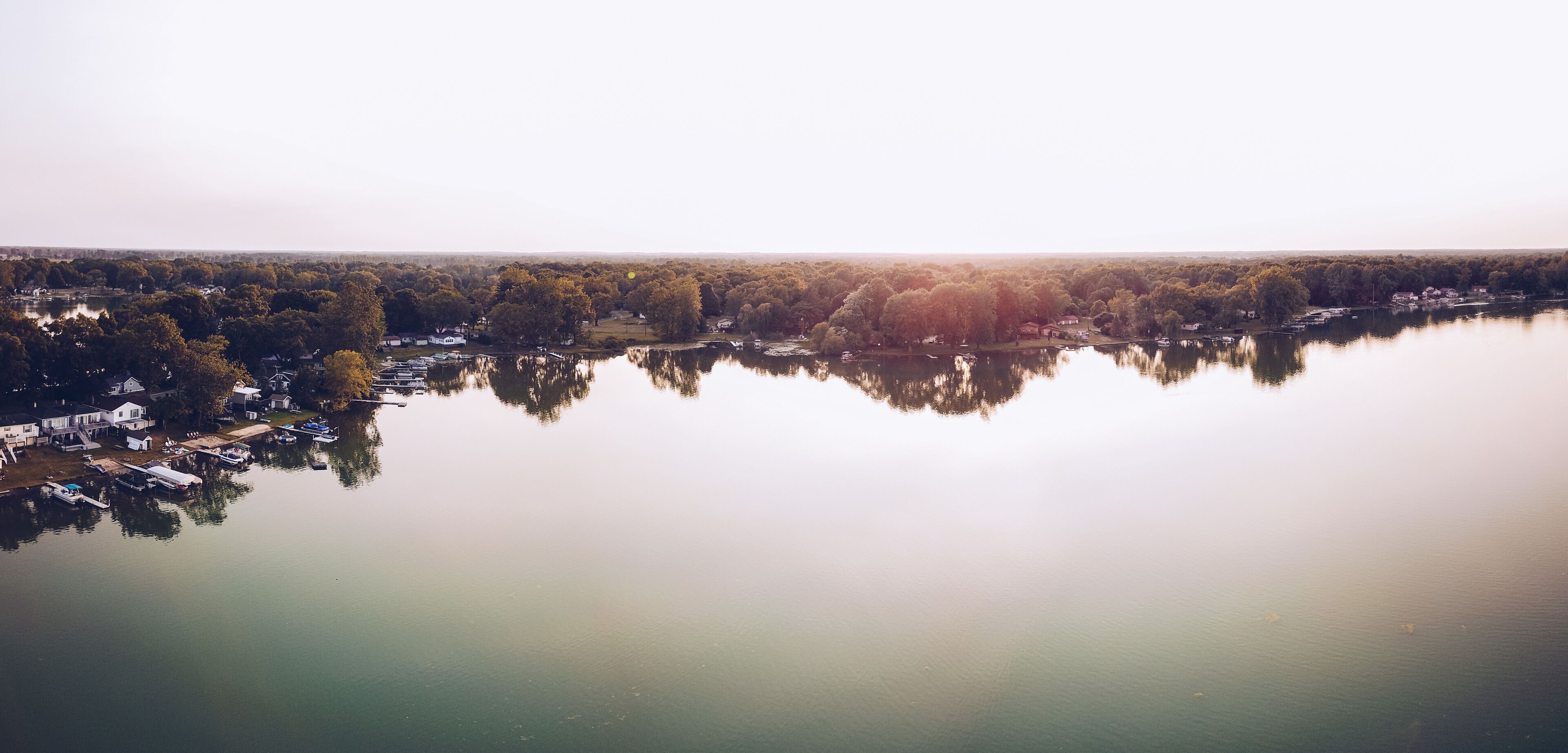 A calm lake with reflections of trees and houses, surrounded by a forested area.