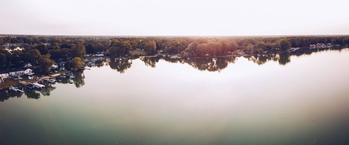 A calm lake with reflections of trees and houses, surrounded by a forested area.