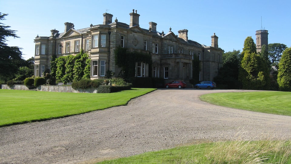 A large, historic stone building with a gravel driveway leading to a red and blue car parked in front.
