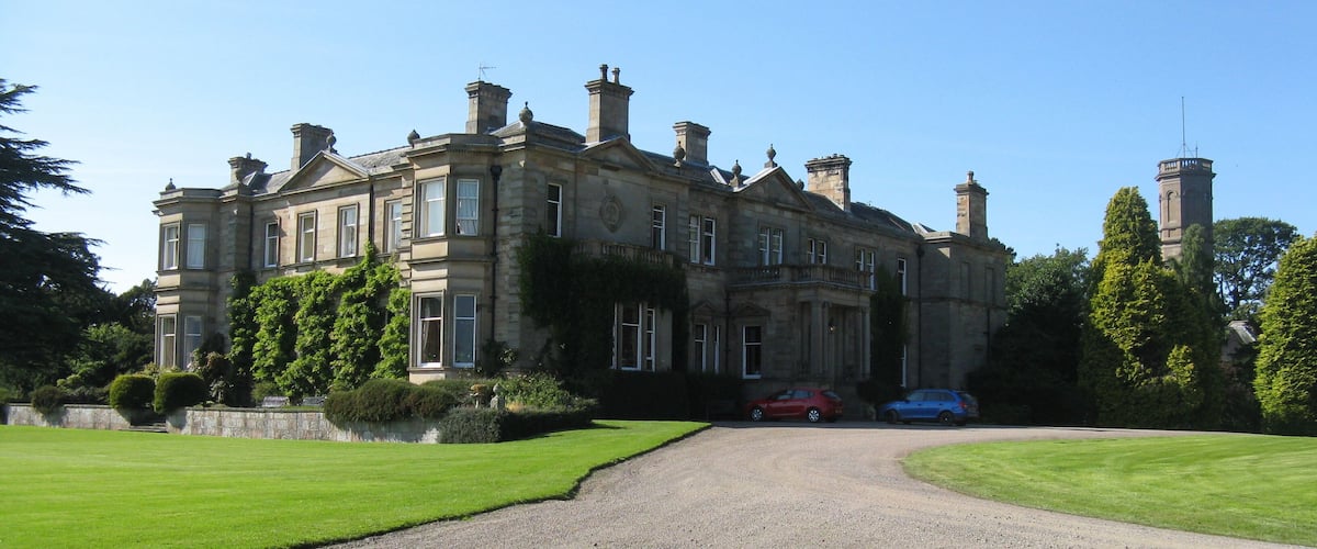 A large, historic stone building with a gravel driveway leading to a red and blue car parked in front.