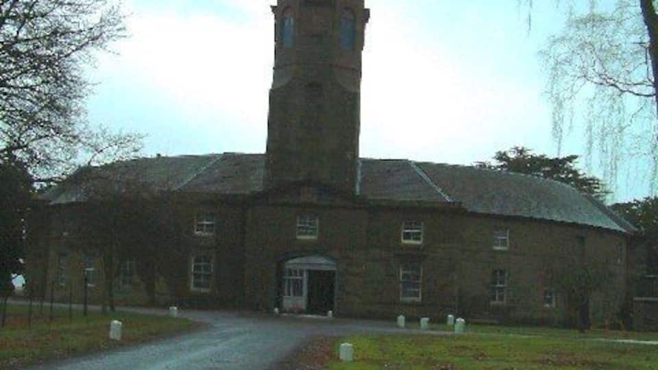 A tall tower attached to a brick building with a paved driveway leading to the entrance.