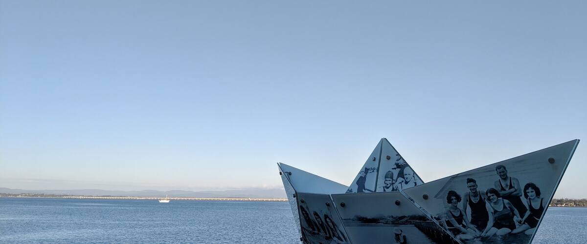 A sailboat with a triangular sail and a black-and-white photo of people on it, set against a calm sea and distant shoreline.