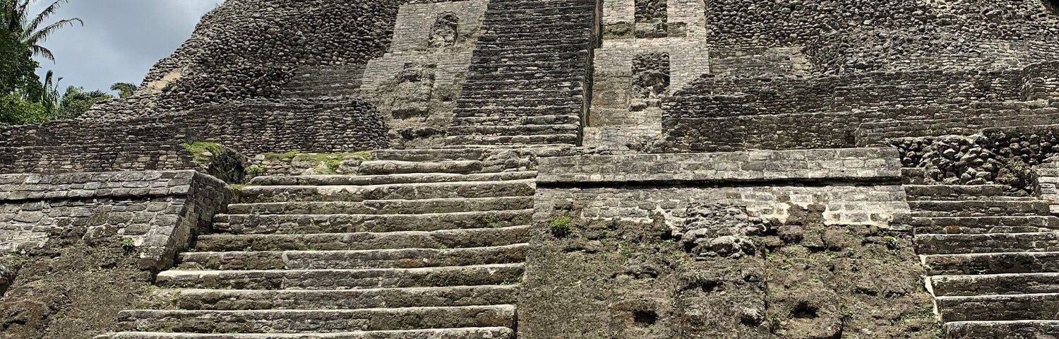 Uma pirâmide de pedra com degraus que levam a um templo, cercada por vegetação e árvores.