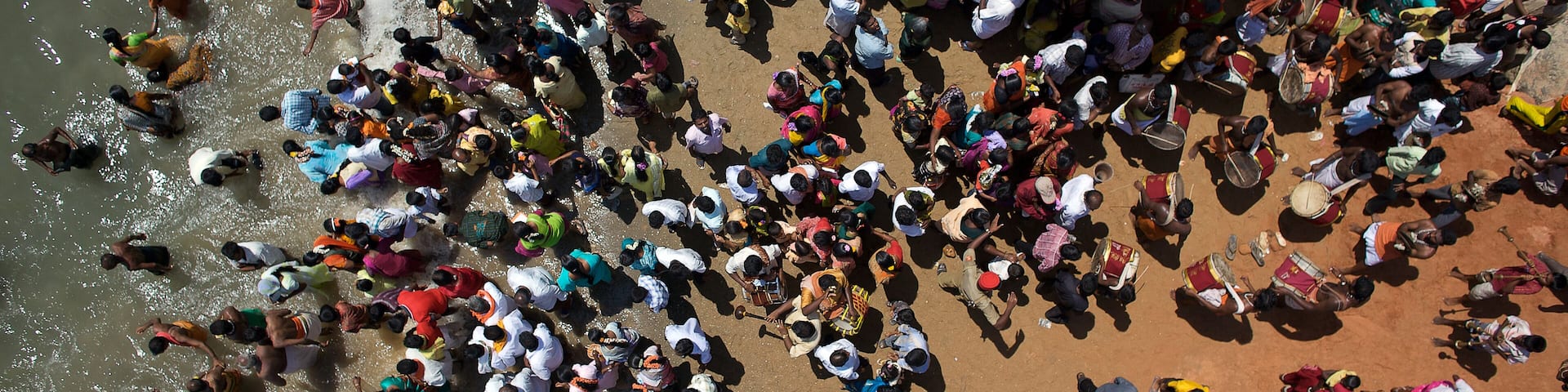 A crowded outdoor scene with people gathered on a reddish-brown surface, possibly a beach or plaza.