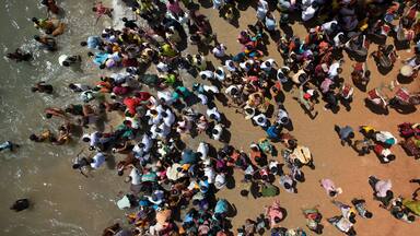 A crowded outdoor scene with people gathered on a reddish-brown surface, possibly a beach or plaza.