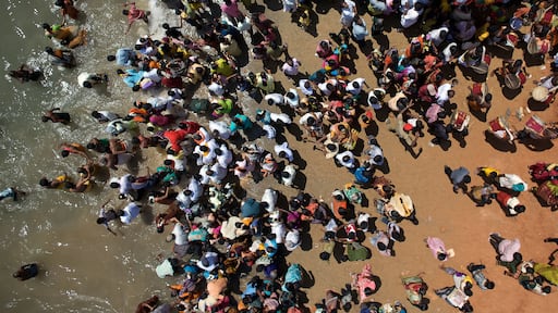 A crowded outdoor scene with people gathered on a reddish-brown surface, possibly a beach or plaza.