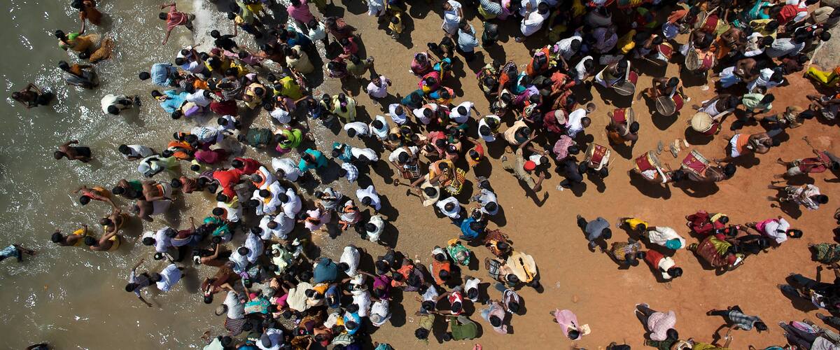 A crowded outdoor scene with people gathered on a reddish-brown surface, possibly a beach or plaza.