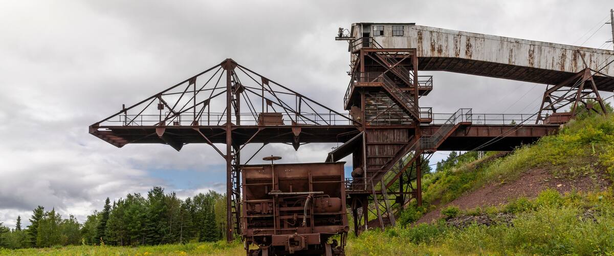 A rusted railway bridge and train tracks in a grassy area with trees in the background.