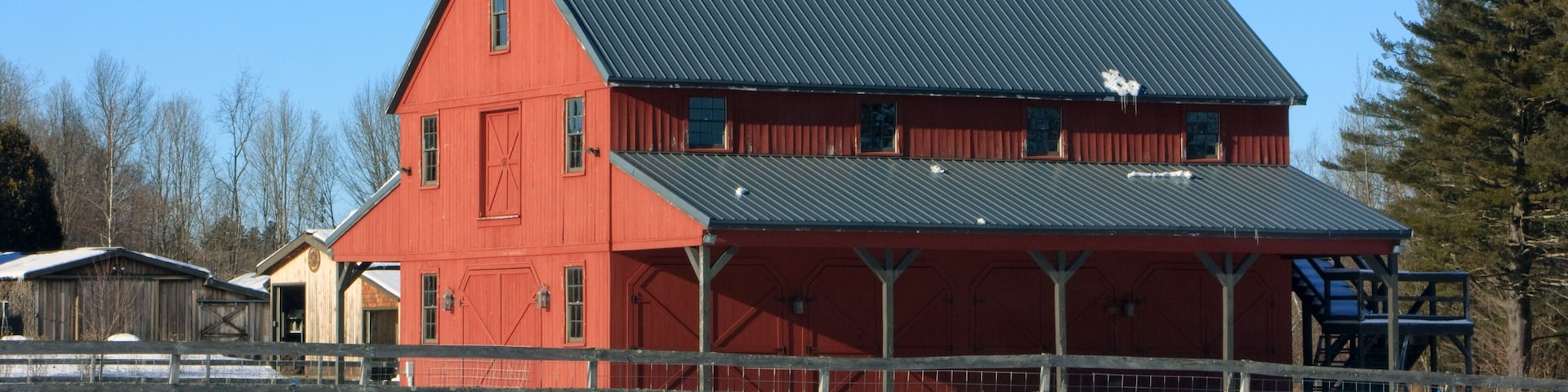 A red barn with a metal roof, surrounded by a snow-covered landscape and a wooden fence.