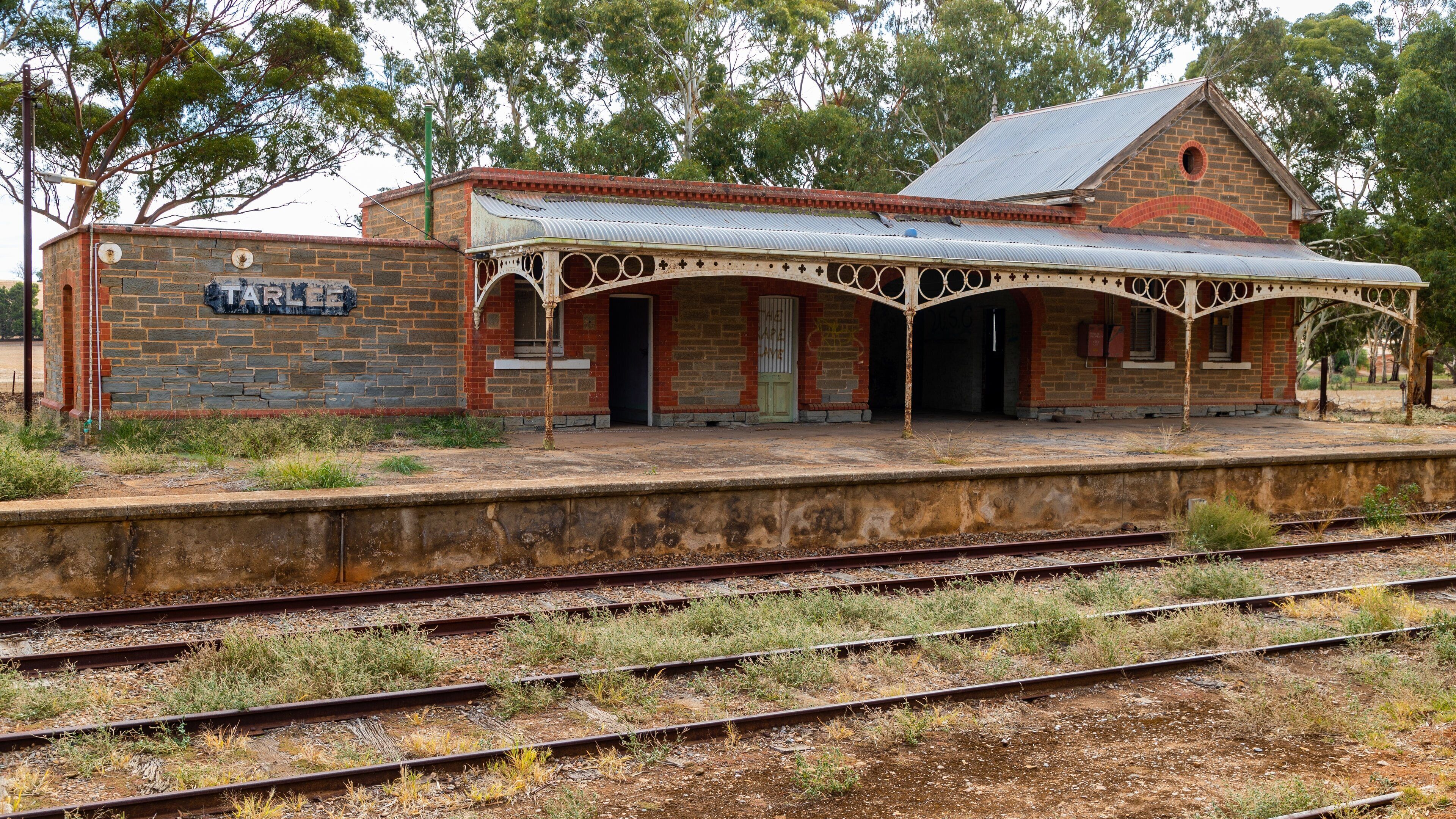 A historic railway station with a stone wall, a covered waiting area, and multiple railway tracks.