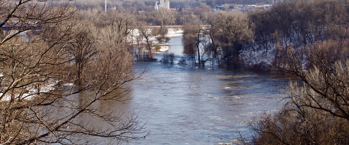A river with a bridge, surrounded by leafless trees and a hilly landscape in the background.