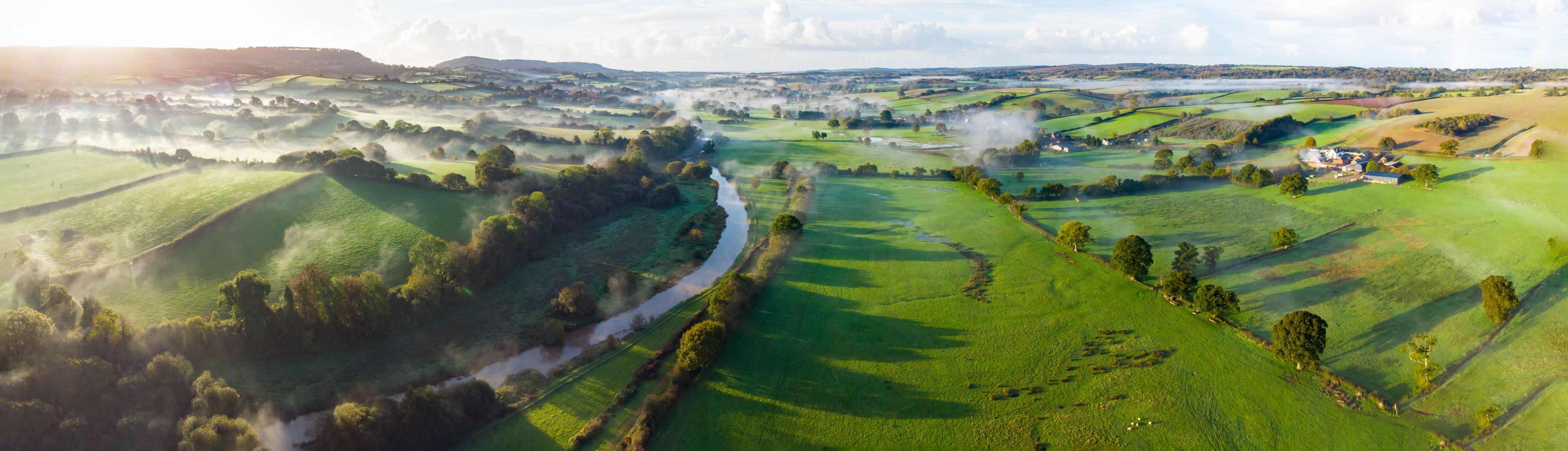 A lush green valley with rolling hills, a river, and a small cluster of buildings.