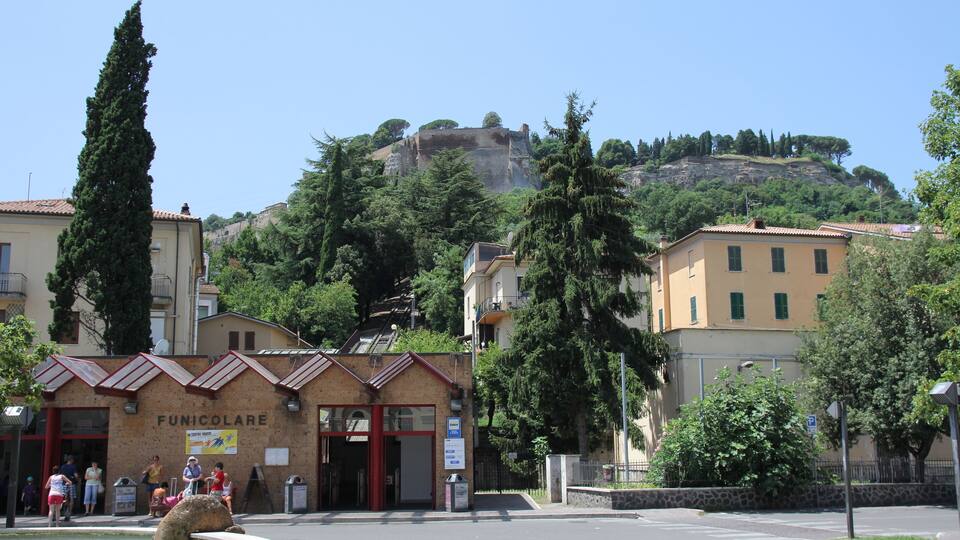 A town square with a building labeled "FUNICOLARE," surrounded by trees and a hill with a castle-like structure in the background.