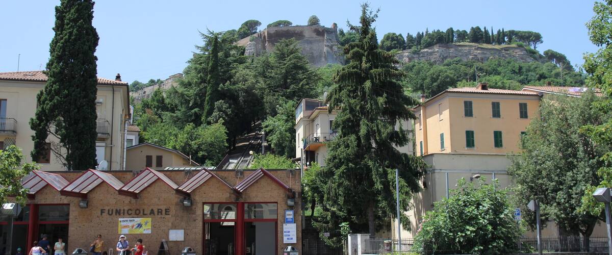 A town square with a building labeled "FUNICOLARE," surrounded by trees and a hill with a castle-like structure in the background.