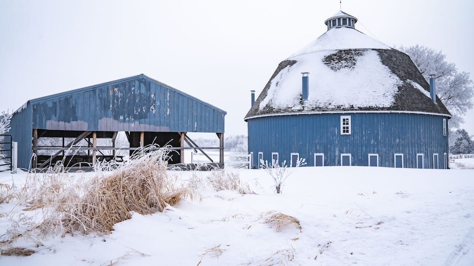 A snow-covered barn and silo in a rural setting.