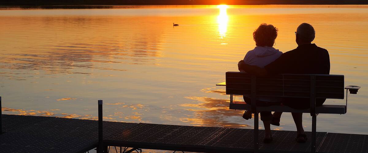 Two people sitting on a bench facing a sunset over a calm lake.