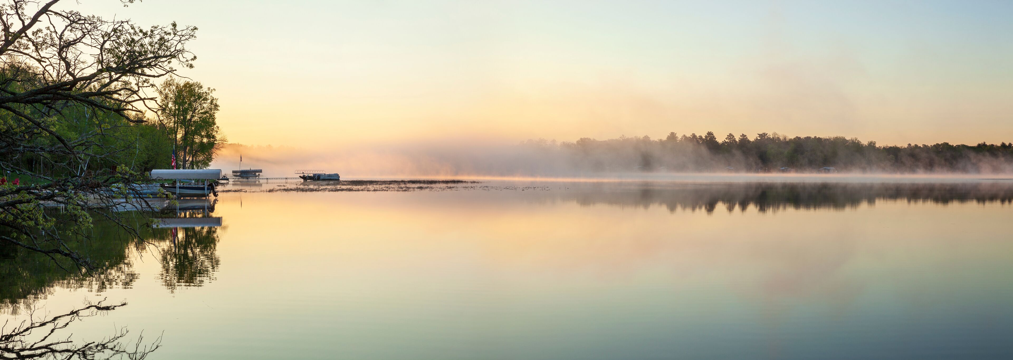 A calm lake with mist rising from the water, reflecting the trees and a dock with boats.