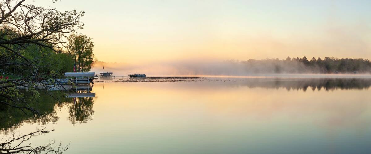 A calm lake with mist rising from the water, reflecting the trees and a dock with boats.
