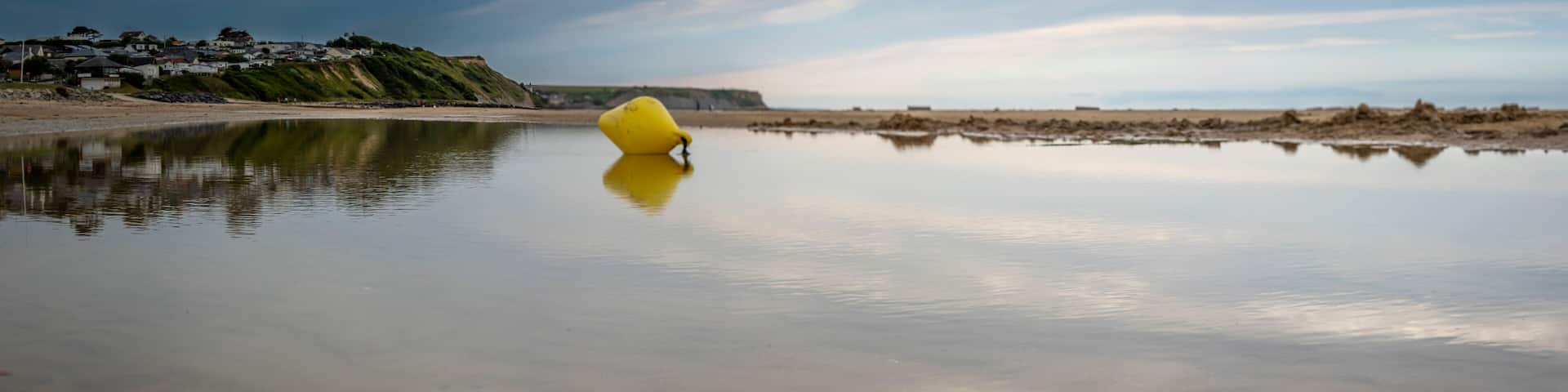 Saint-Come-de-Fresne, France - 08 08 2025: Panoramic view of a yellow buoy reflecting on the water, the sea, the beach, the cliff and the city with cloudy sky