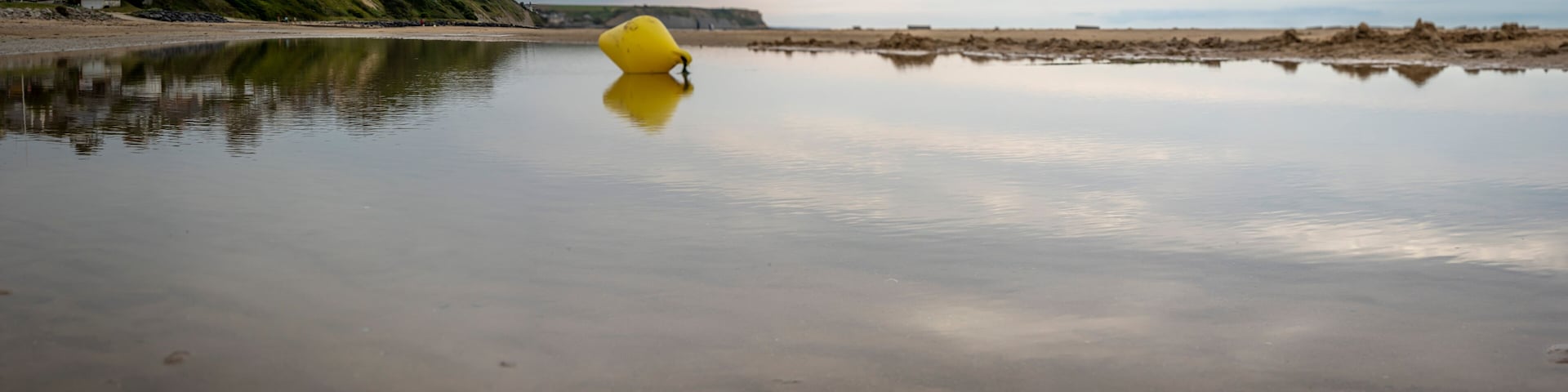 Saint-Come-de-Fresne, France - 08 08 2025: Panoramic view of a yellow buoy reflecting on the water, the sea, the beach, the cliff and the city with cloudy sky