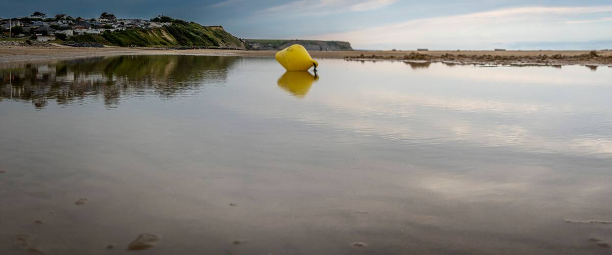 A calm body of water with a yellow buoy in the center, a sandy beach, and a hill with houses in the background.