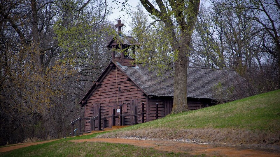 A wooden church with a bell tower, surrounded by trees and a grassy hill.