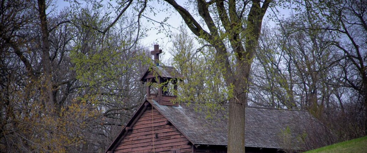 A wooden church with a bell tower, surrounded by trees and a grassy hill.