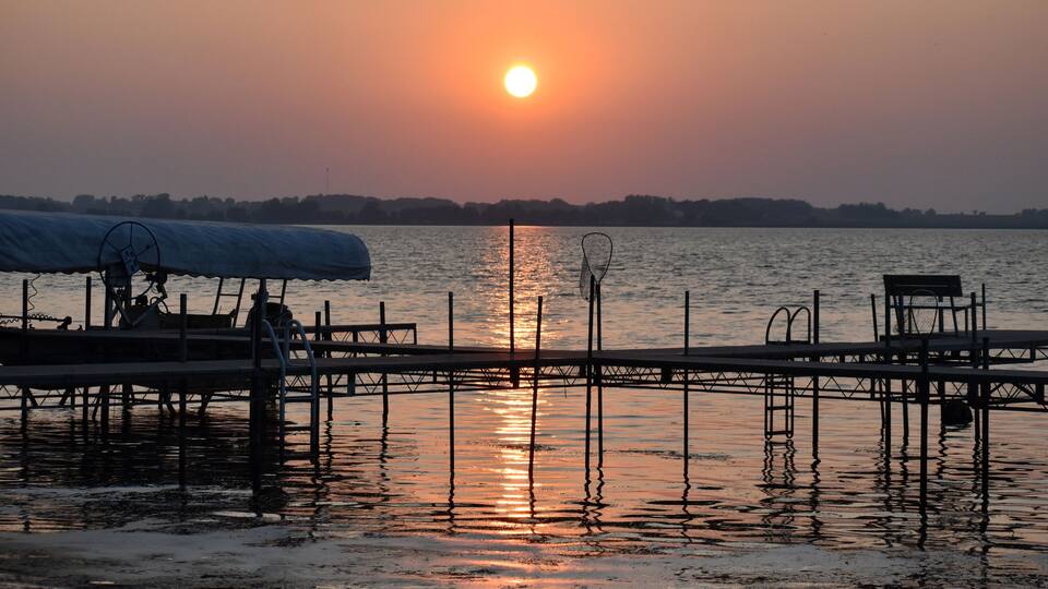 A sunset over a calm lake with a dock and chairs.