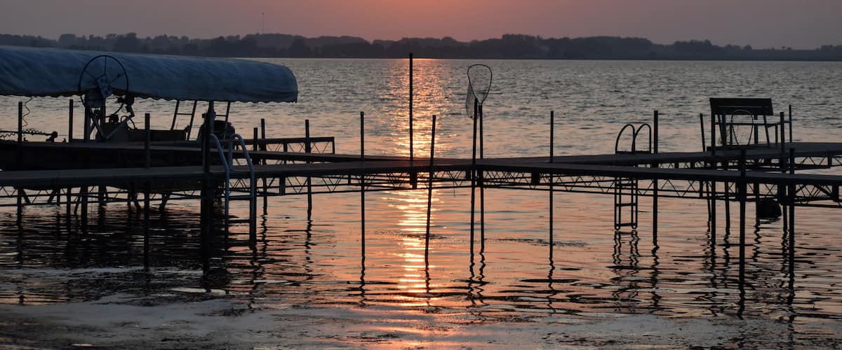 A sunset over a calm lake with a dock and chairs.