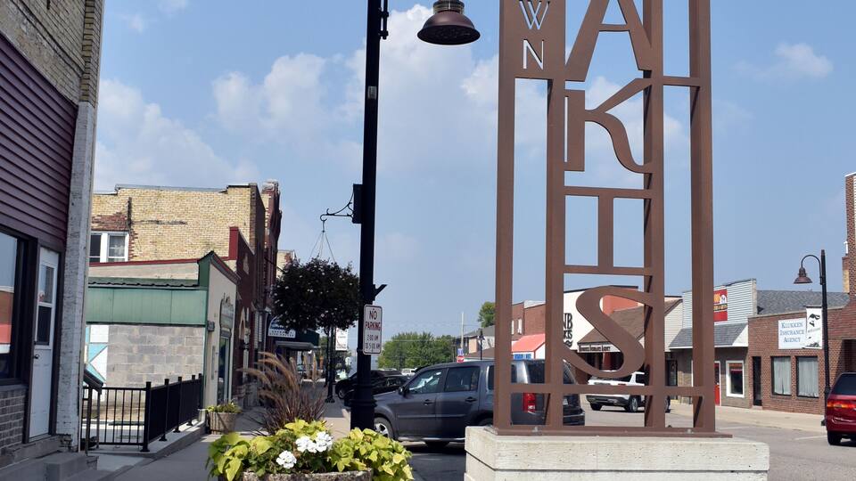A metal sign with 'DOWNTOWN' and 'OSAKIS' on it, a street lamp, a planter with flowers, and a building in the background.