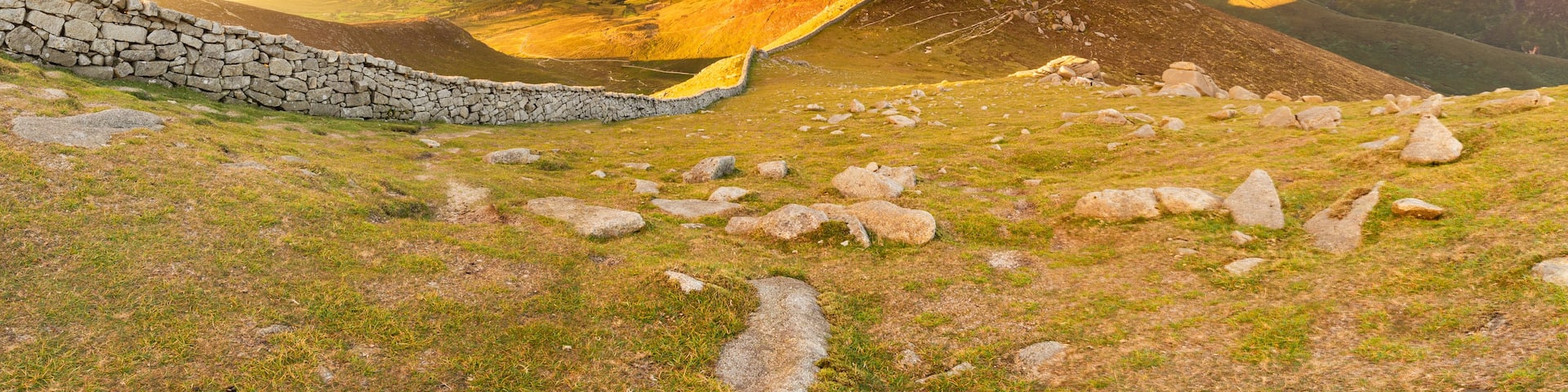 A stone wall, rocky terrain, and a mountainous landscape with a cloudy sky.