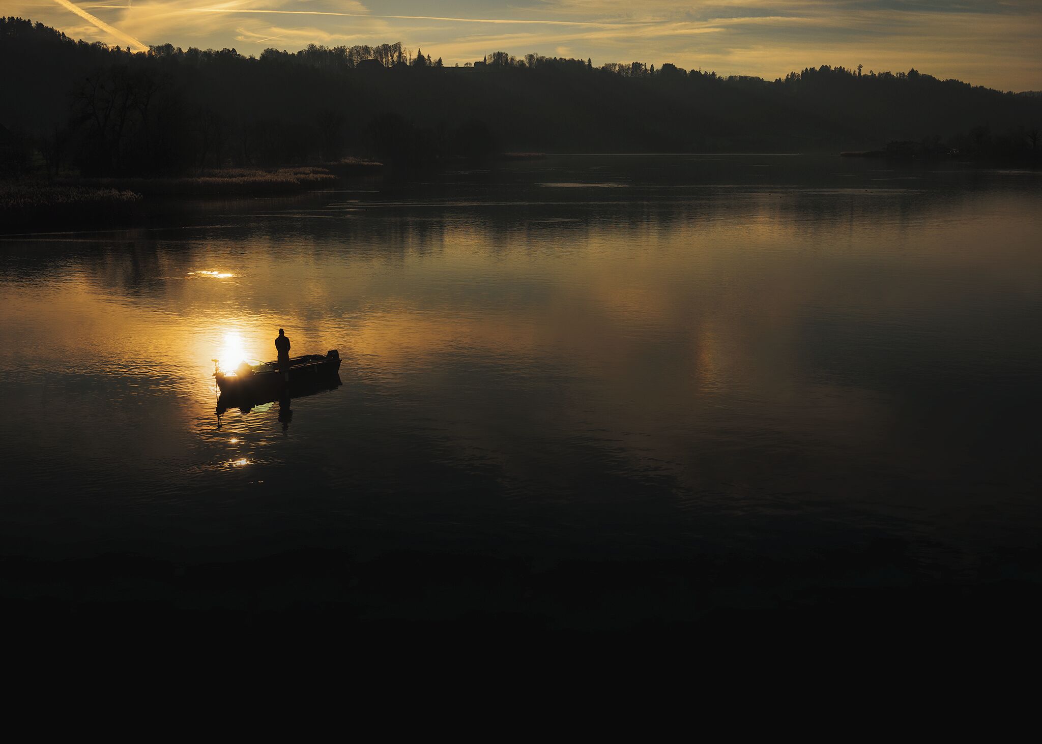 A calm lake at sunrise with a boat and a person silhouetted against the sun.