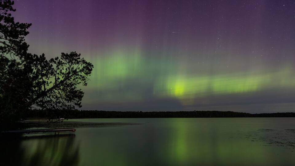 A night sky with the Northern Lights over a calm lake with a dock and trees.