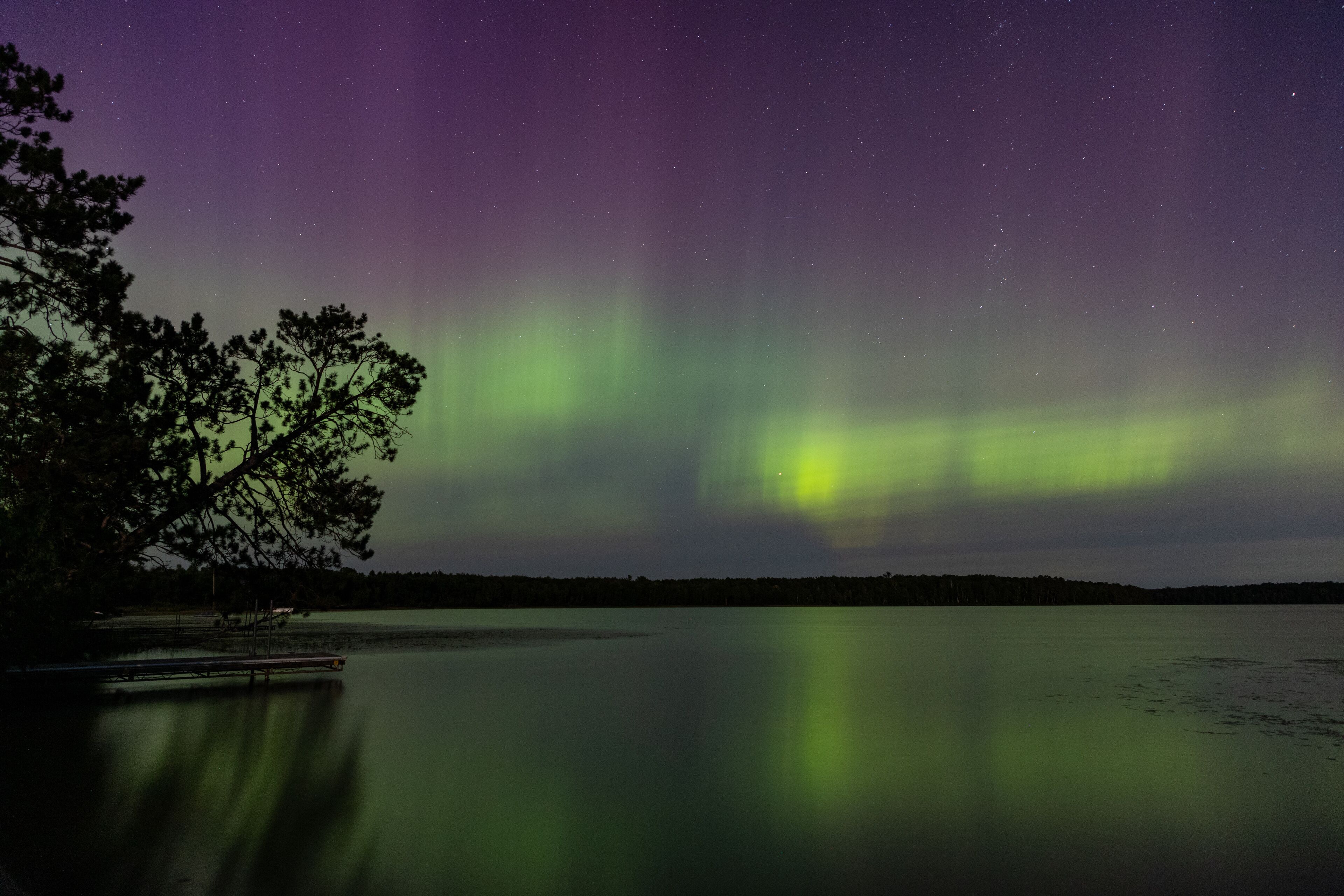 A night sky with the Northern Lights over a calm lake with a dock and trees.