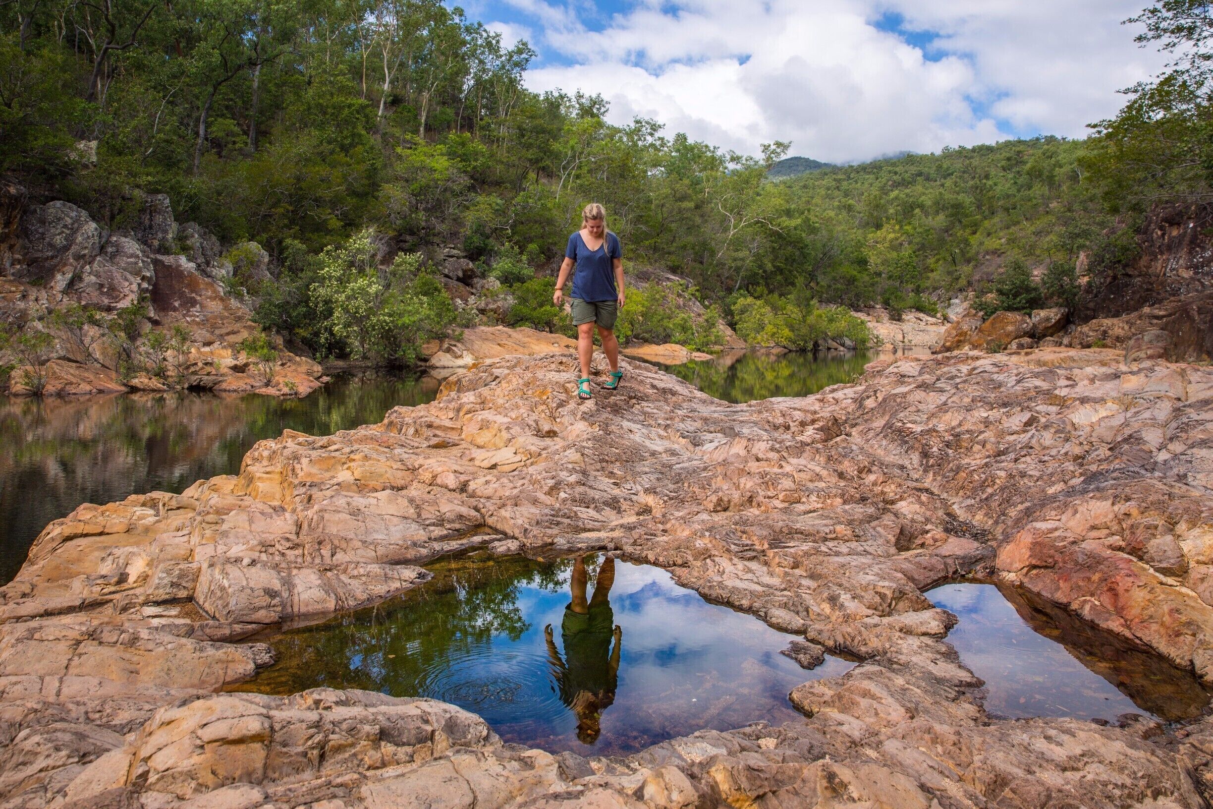 A person standing on rocky terrain with a reflective water pool surrounded by trees and hills.