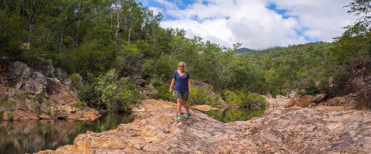 A person standing on rocky terrain with a reflective water pool surrounded by trees and hills.