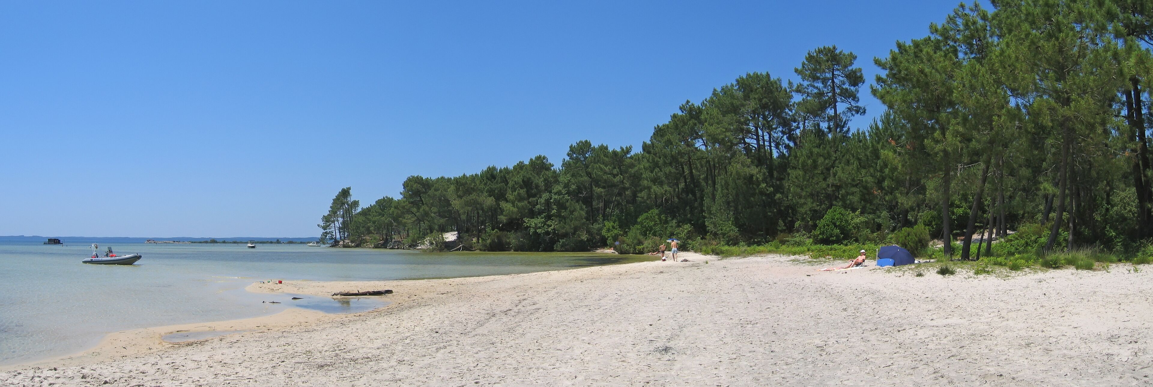 A beach with clear water, a boat, and a forest in the background.
