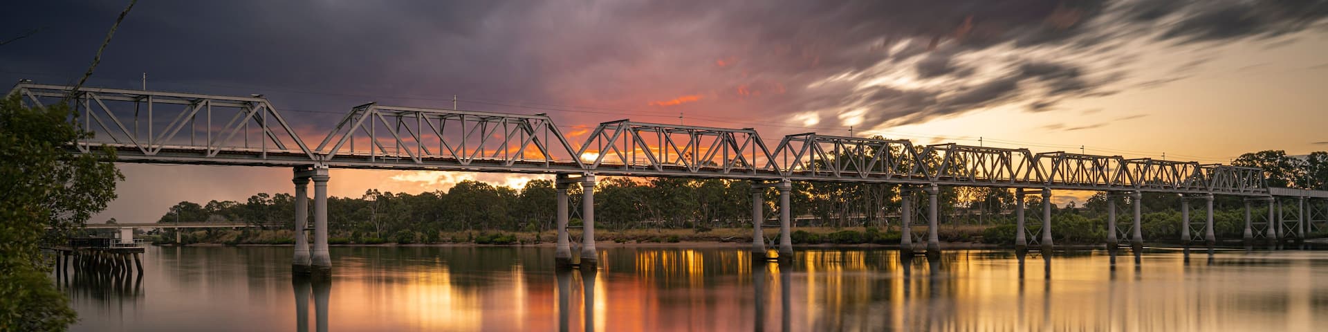 A steel bridge over calm water with a sunset reflection.