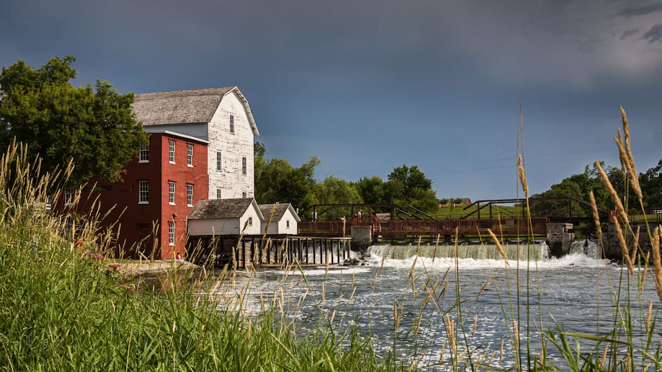 A red and white mill by a river with a wooden bridge and lush greenery.