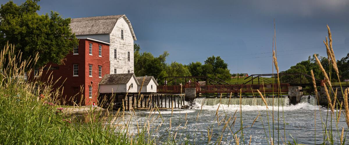 A red and white mill by a river with a wooden bridge and lush greenery.