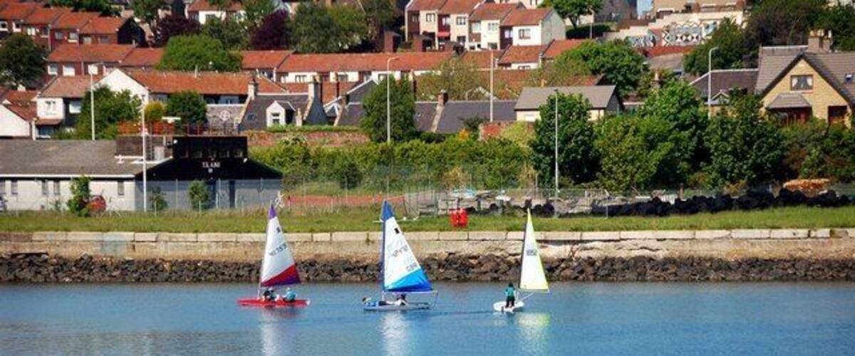 Three sailboats on a calm body of water with houses in the background.