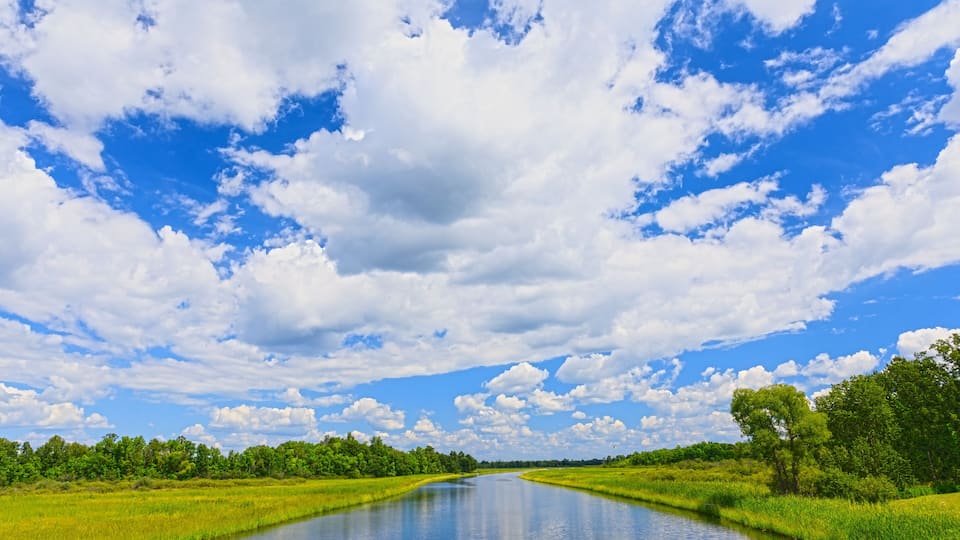 A river flanked by lush green fields and a clear blue sky with scattered clouds.