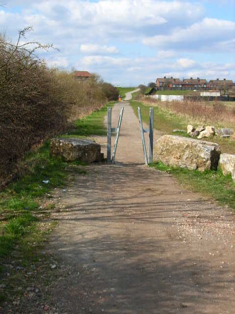 A rural pathway leading to a gated entrance with a stone wall on one side and a residential area in the background.